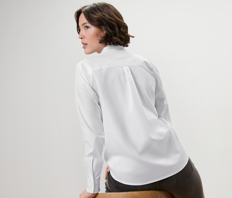 Kobieta siedzi bokiem na drewnianym krześle, ma na sobie białą bluzkę z falbankami.