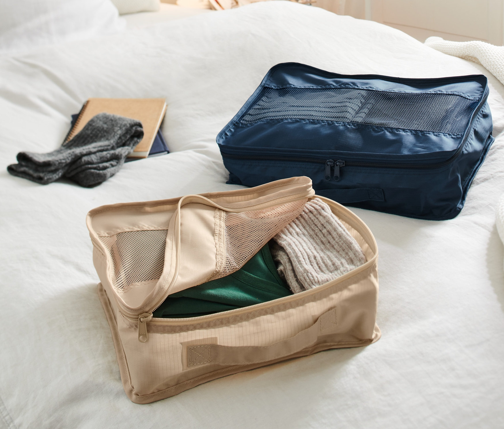 Dwa organizery podróżne leżą na łóżku. Beżowy organizer jest otwarty i wypełniony ubraniami, a granatowy organizer jest zamknięty.