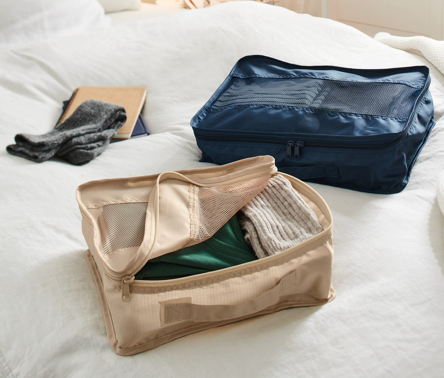Dwa organizery podróżne leżą na łóżku. Beżowy organizer jest otwarty i wypełniony ubraniami, a granatowy organizer jest zamknięty.