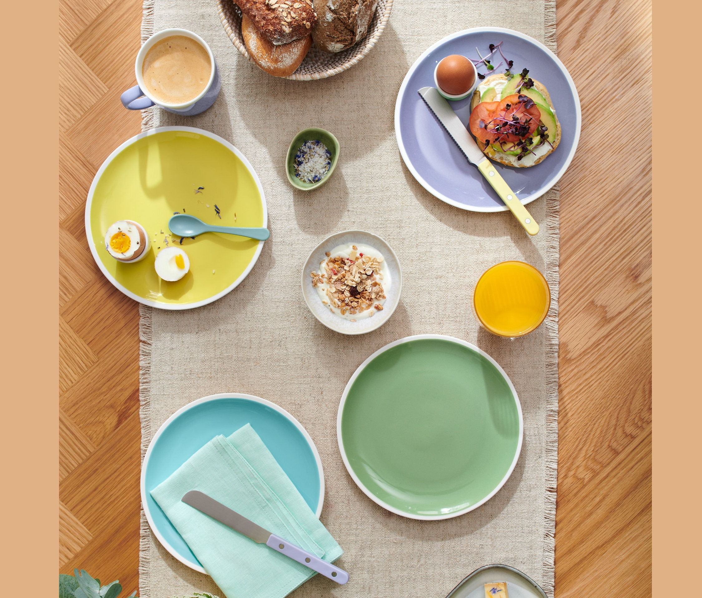 Widok z góry na nakryty stół z talerzami śniadaniowymi (4 sztuki), filiżanką kawy, sokiem, pieczywem w koszyku i talerzem serów.