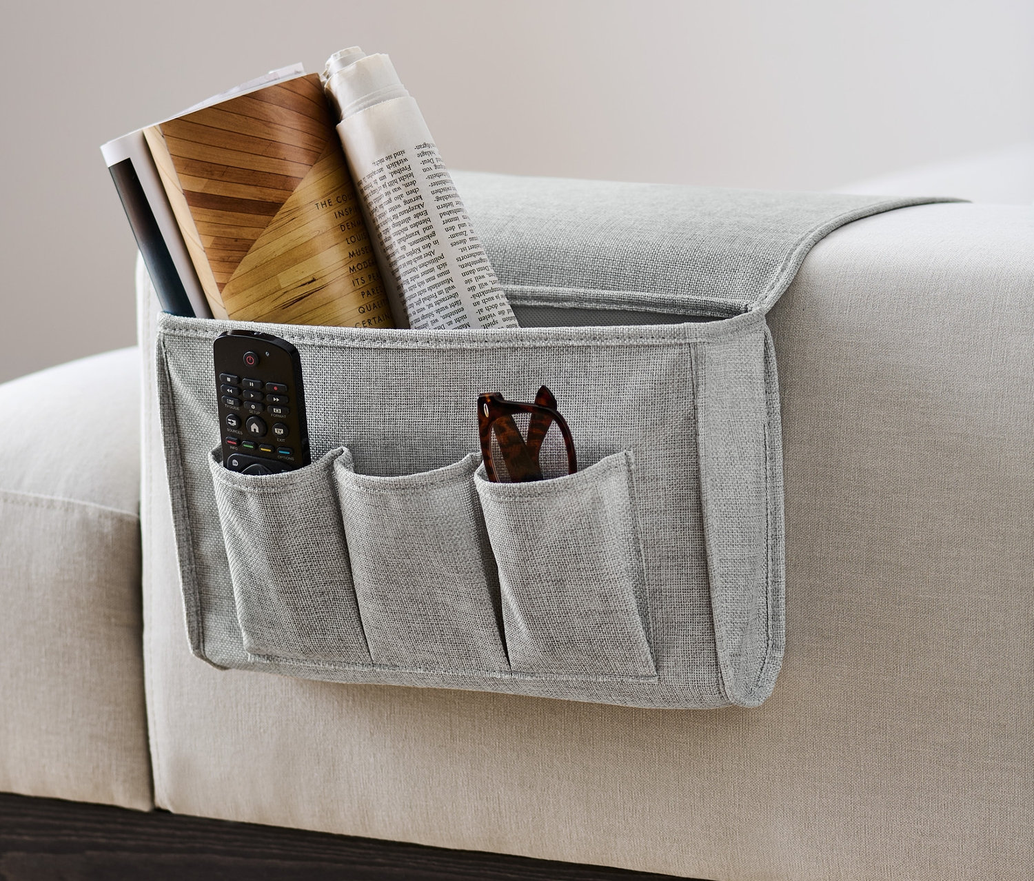Szary organizer na łóżko i kanapę wisi z boku sofy i zawiera pilot, okulary, książki i gazetę.