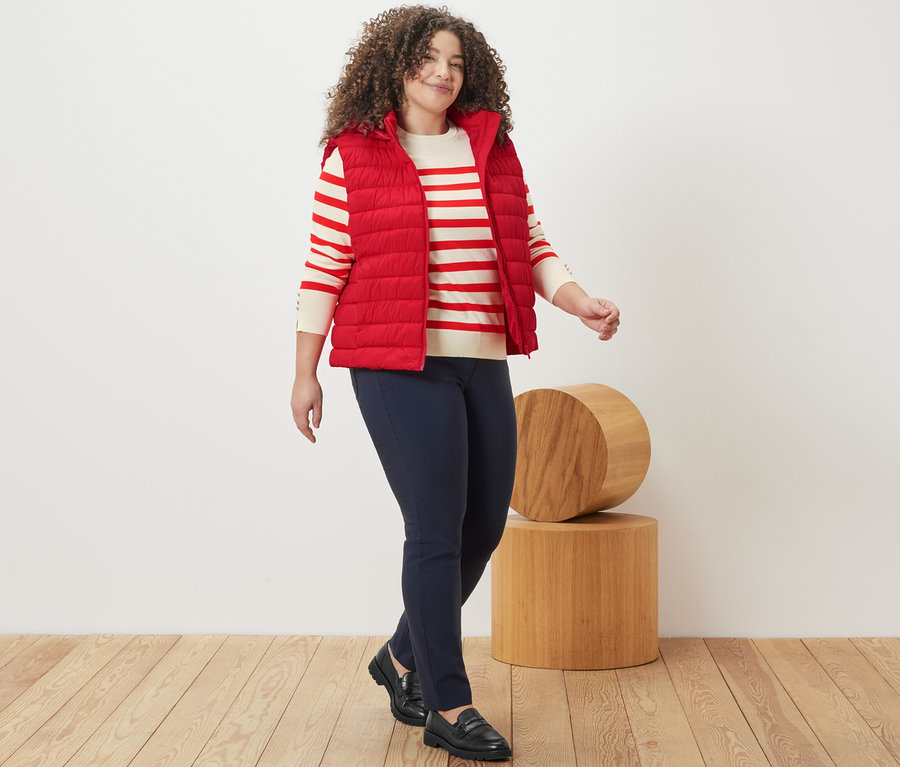 Kobieta idzie, ma na sobie czerwony cienki sweter w paski, kamizelkę pikowaną z kapturem i granatowe spodnie z bengaliny.
