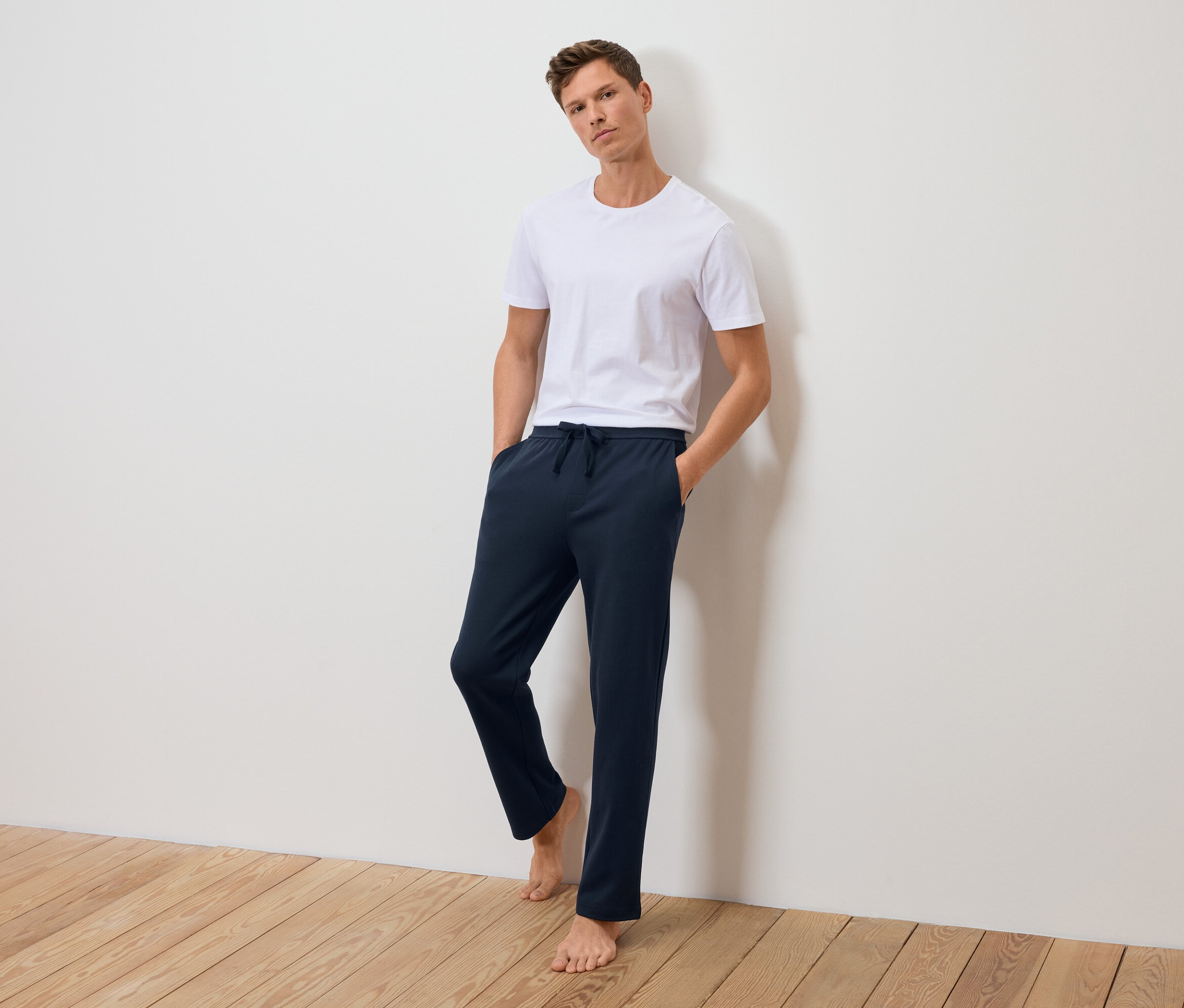 Mężczyzna stoi oparty o ścianę w białej koszulce i granatowych spodniach dresowych.