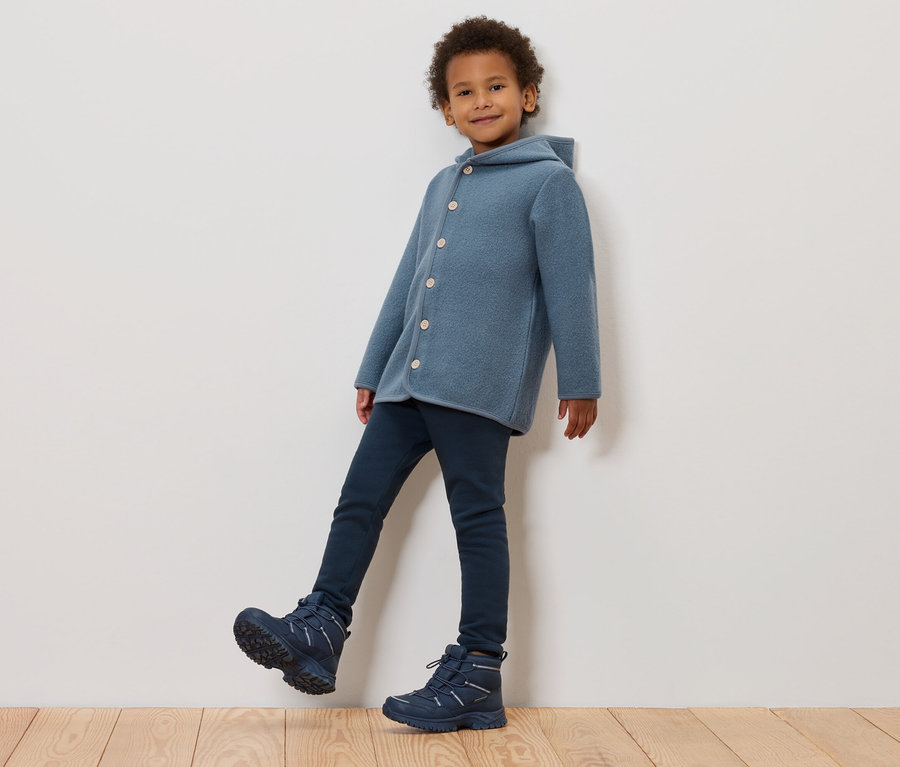 Chłopiec w niebieskim wełnianym płaszczu z kapturem, ciemnoniebieskich dżinsach i niebieskich zimowych butach pozuje pod ścianą.