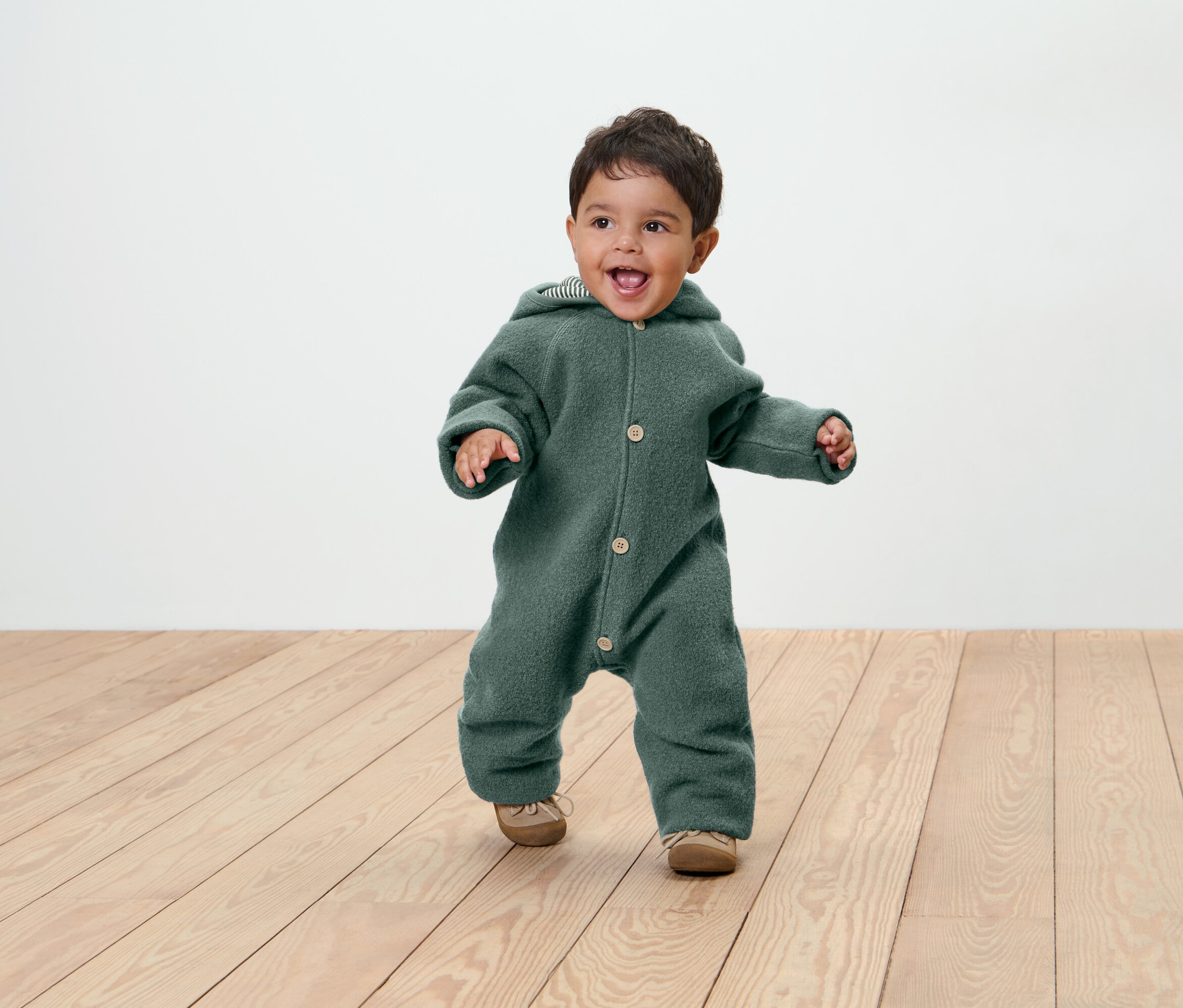 Uśmiechnięte dziecko w zielonym kombinezonie z kapturem i butach na drewnianej podłodze.