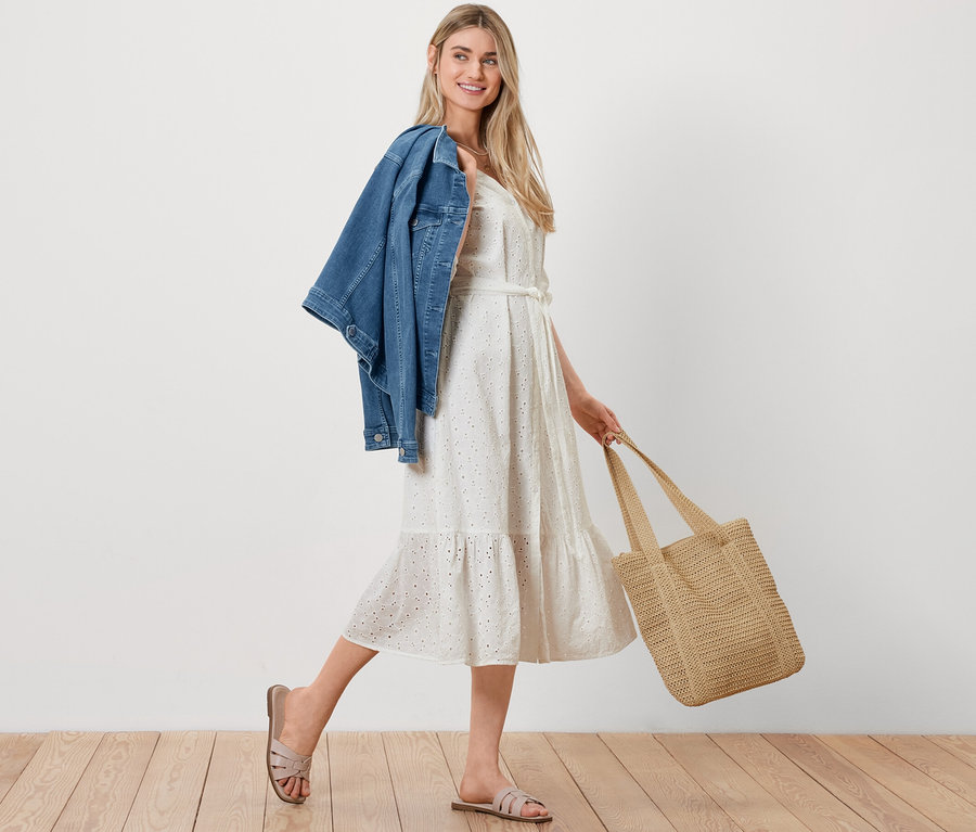 Kobieta idzie z kurtką dżinsową przewieszoną przez ramię, torbą shopperką z plecionki i sukienką koszulową z haftem ażurowym.