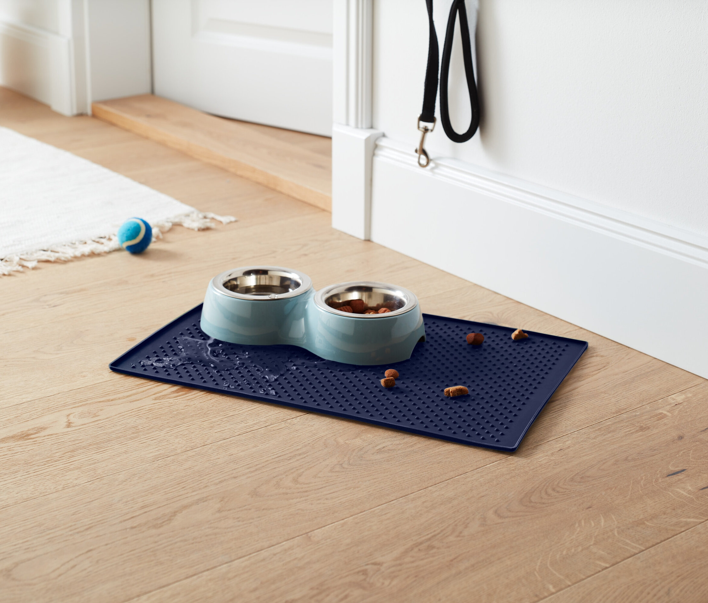 Niebieska podkładka z miskami dla psów, jedna miska pełna jedzenia, na podkładce i wokół niej leżą granulki. W tle biały dywan, niebieska piłka i czarna smycz.