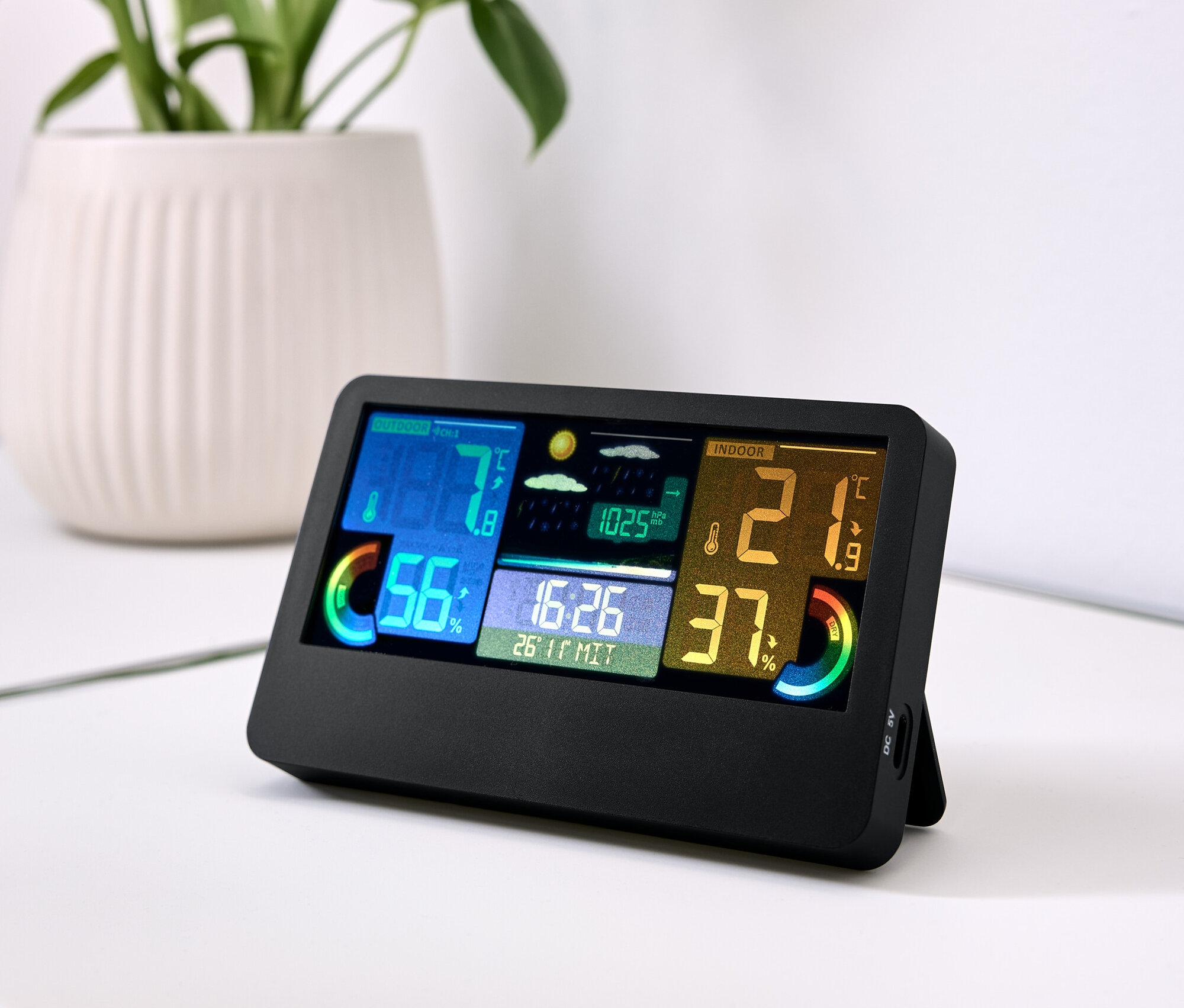 Czarna stacja pogodowa z kolorowym wyświetlaczem pokazuje temperaturę, wilgotność i czas. Obok leżą klucze, a w tle roślina w białej doniczce.