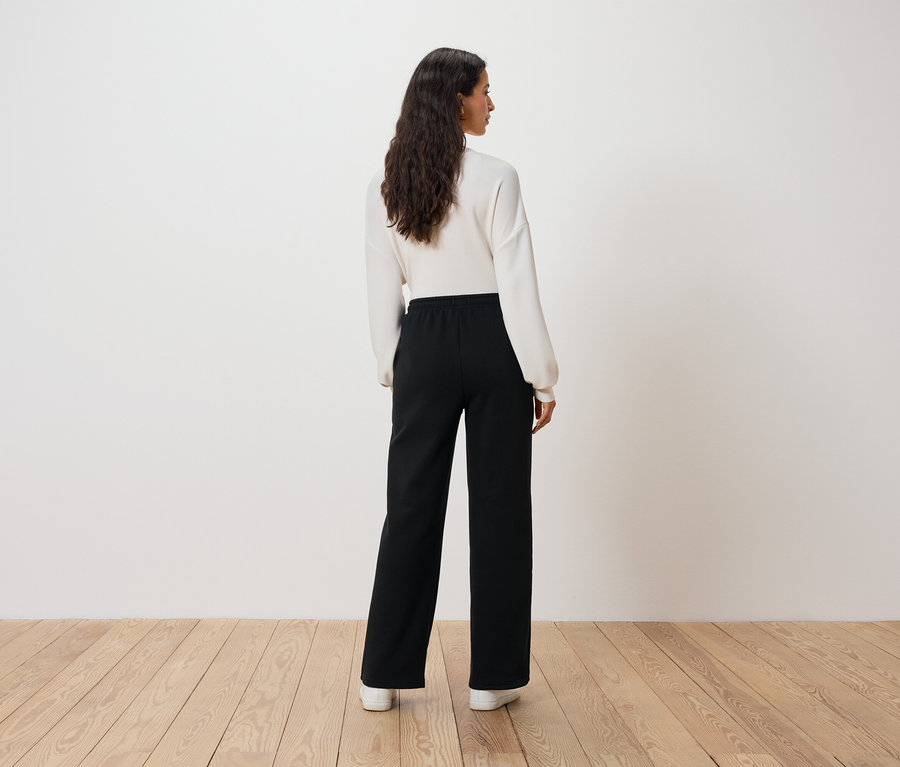 Kobieta w białym swetrze i czarnych spodniach stoi tyłem do widza.