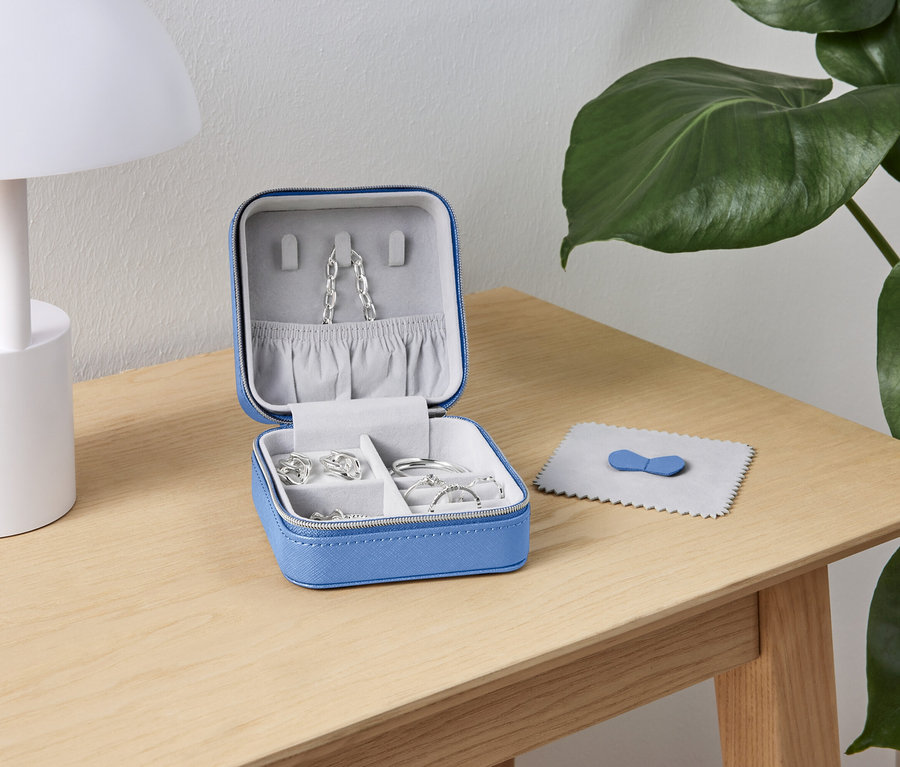 Otwarty, niebieski kuferek na biżuterię ze srebrną biżuterią stoi na drewnianym stole obok białej lampy i zielonej rośliny.