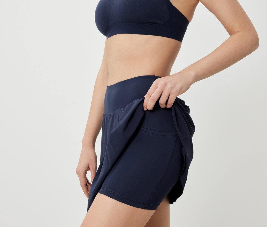 Kobieta pozuje w staniku sportowym i granatowej spódnicy sportowej.