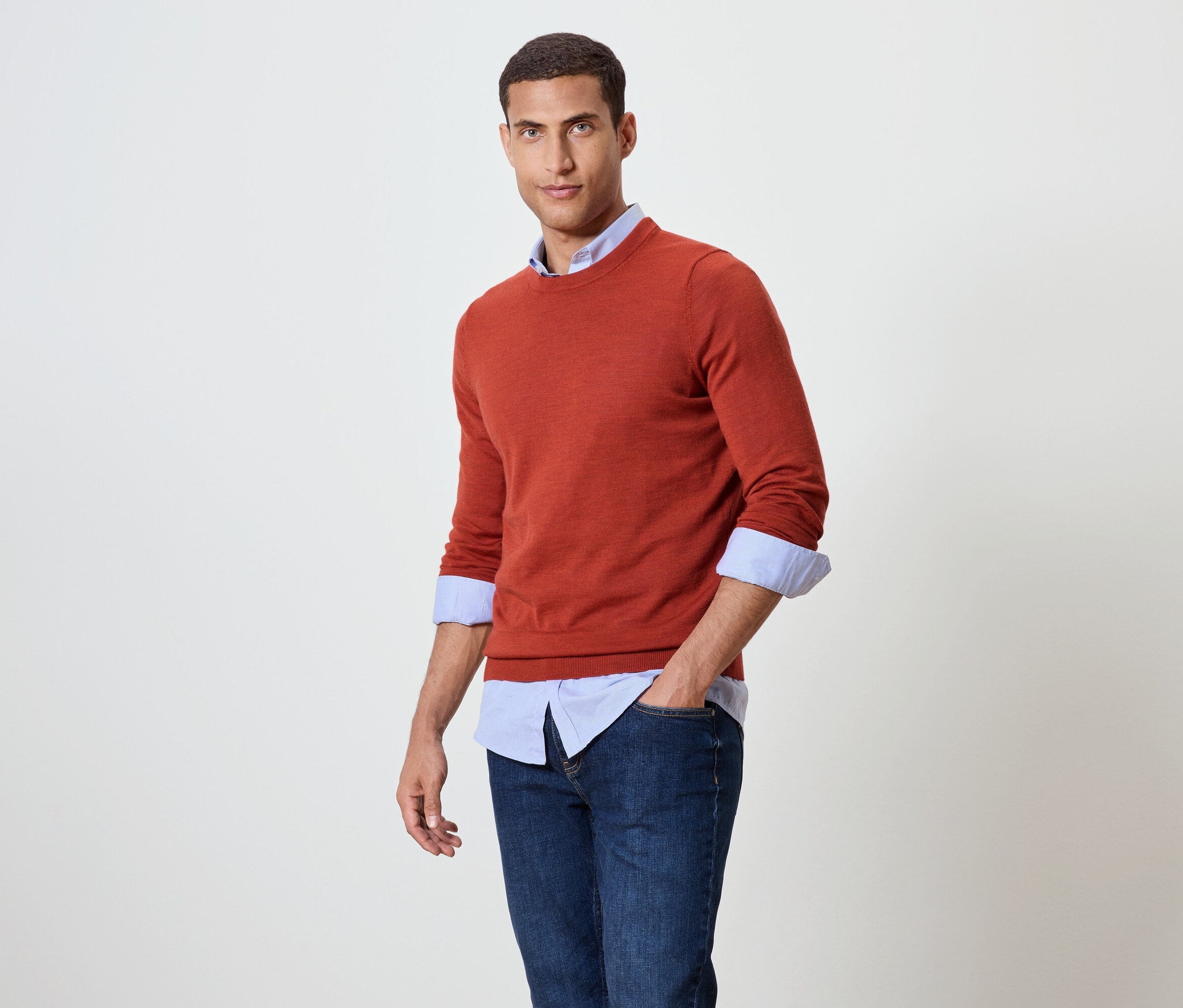 Mężczyzna pozuje w pomarańczowym swetrze nałożonym na niebieską koszulę i dżinsy.
