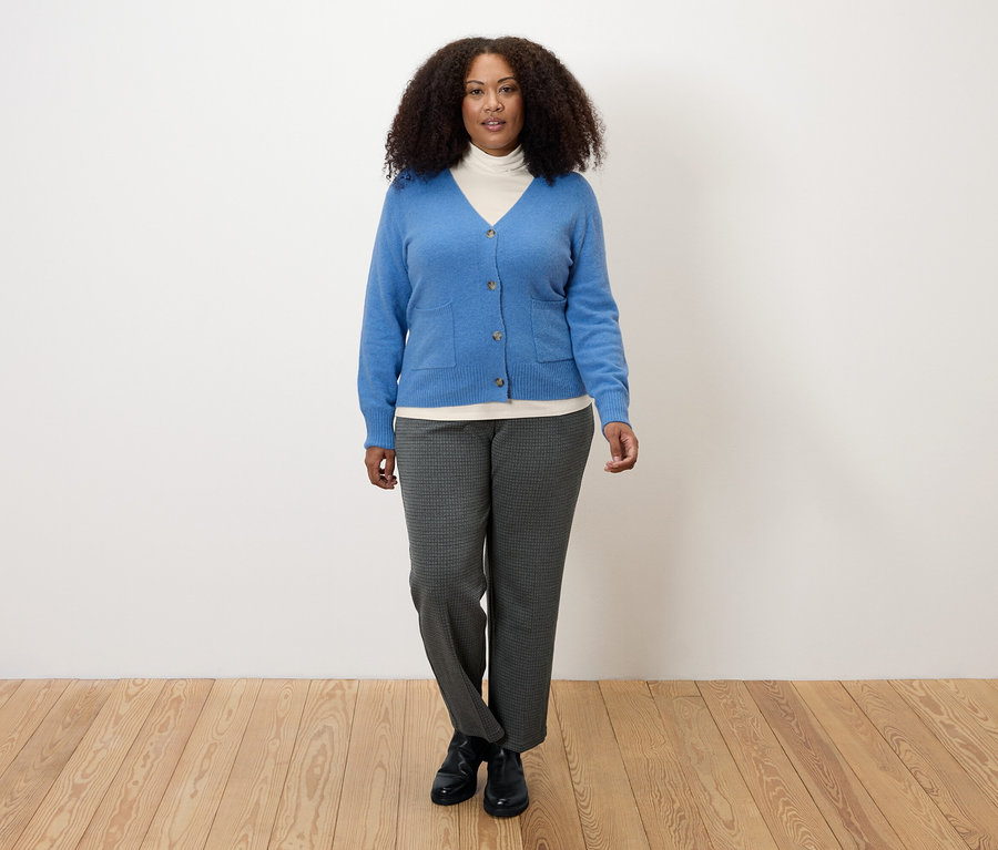 Kobieta w kremowo-białej bluzce z kołnierzem golfowym, niebieskim kardiganie i szarych spodniach.