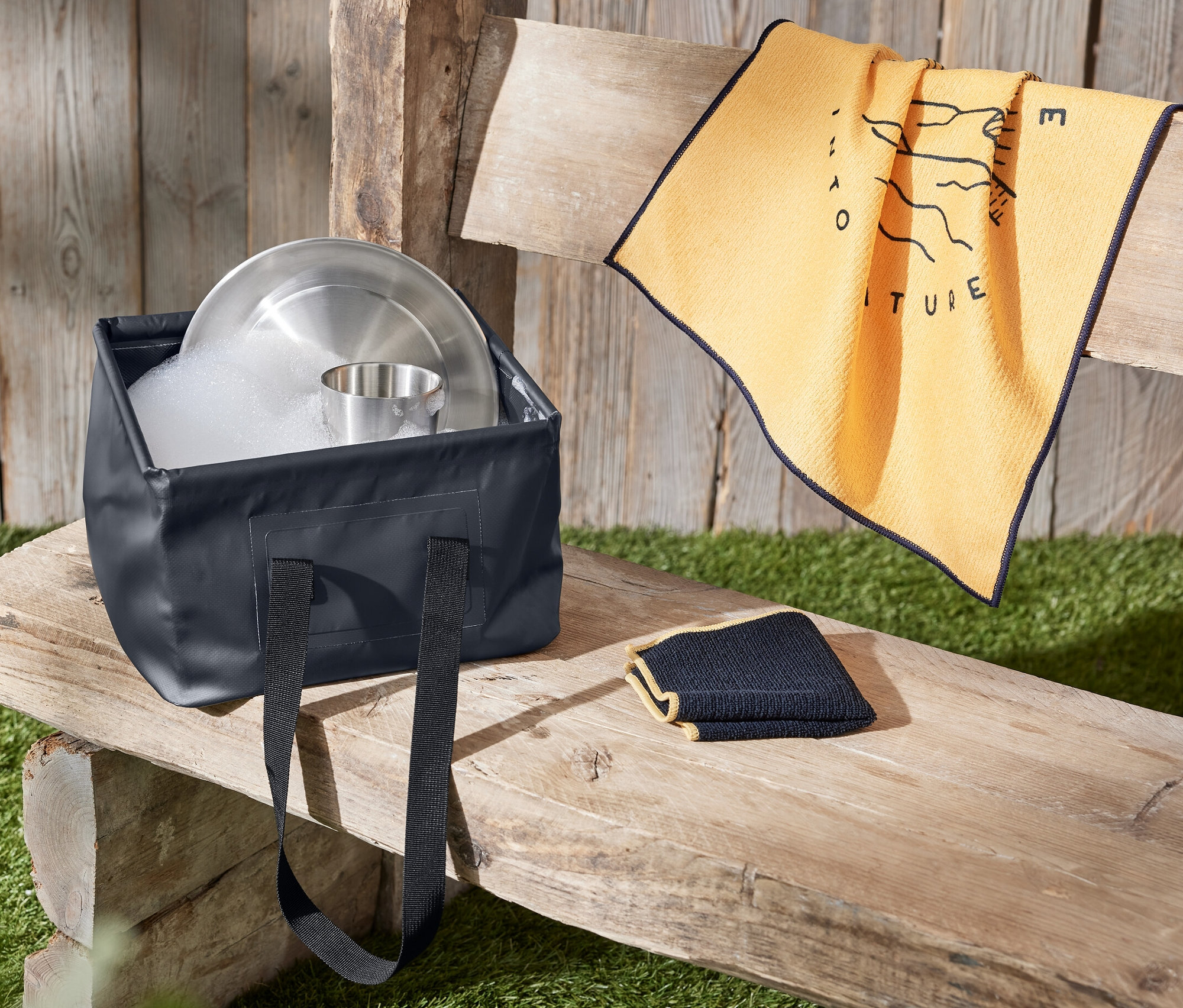 Składana miska do zmywania pełna wody i naczyń stoi na drewnianej ławce obok ręcznika.