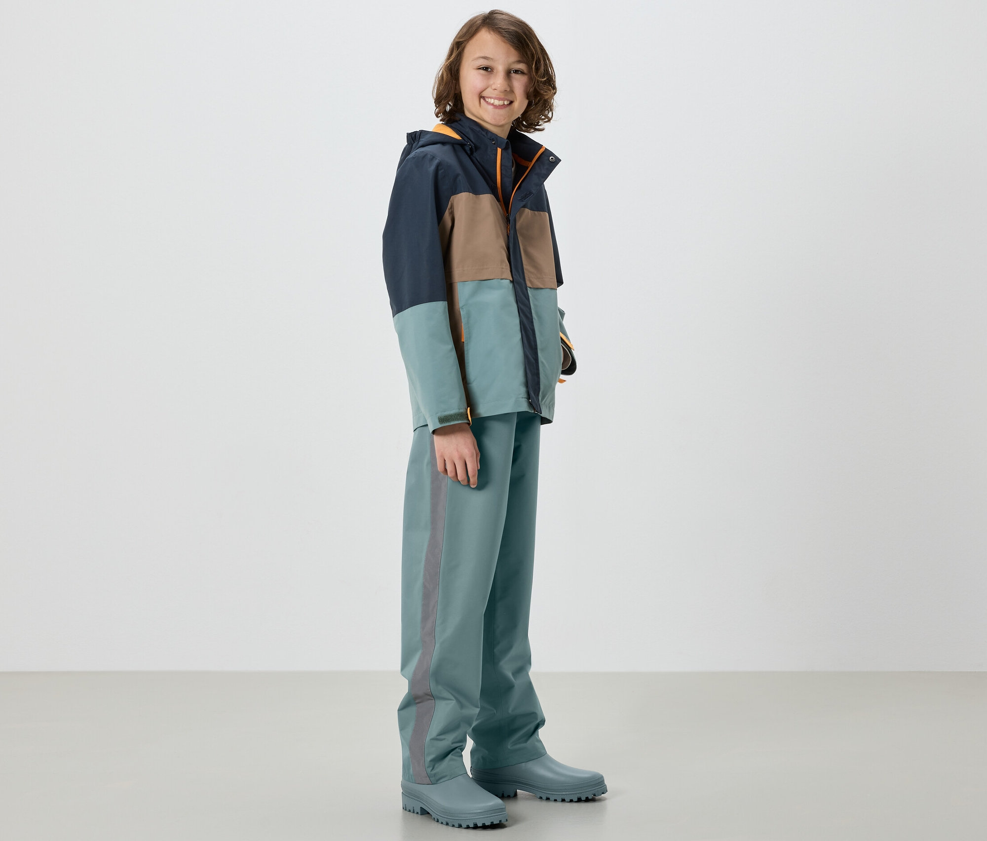 Chłopiec w kurtce i dziecięcych spodniach przeciwdeszczowych, szałwiowych, stoi w kaloszach.