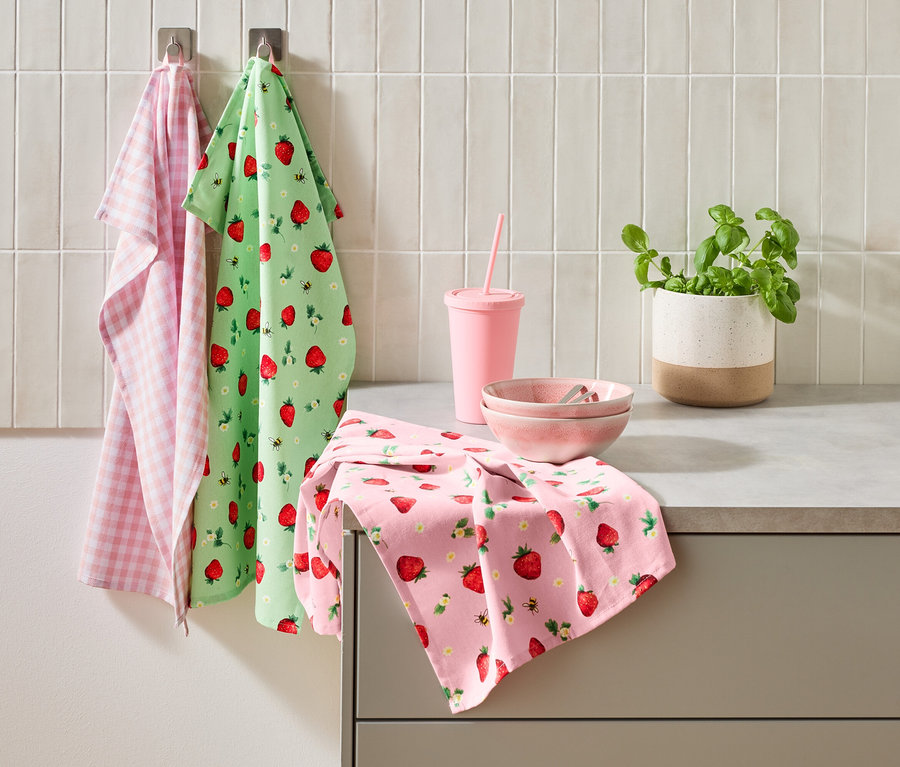 Trzy ściereczki kuchenne, różowy kubek ze słomką, dwie różowe miski i doniczka bazylii stoją na blacie kuchennym.