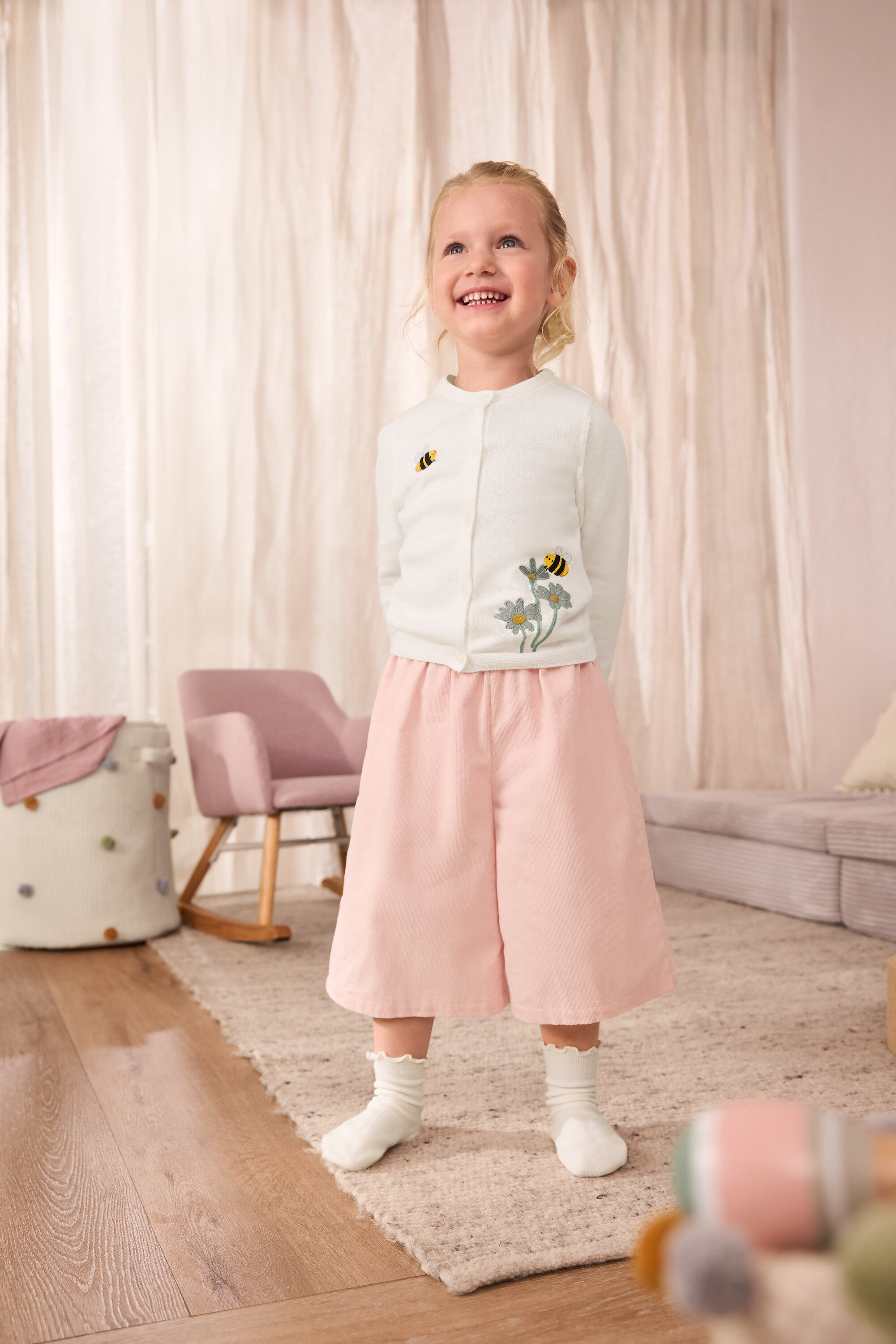 Uśmiechnięta dziewczynka ma na sobie rozpinany sweterek dziecięcy z haftowanymi pszczołami,dziecięce spodnie culotte z bawełny oraz skarpetki z falbanką, 5 par.