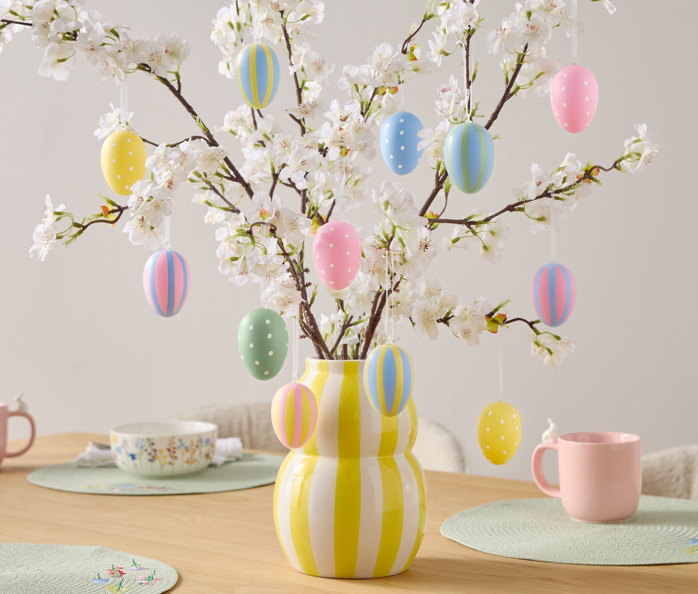 Dekoracyjny stół z pisankami na kwitnących gałęziach w żółto-białym wazonie w paski, różowe kubki i miska.