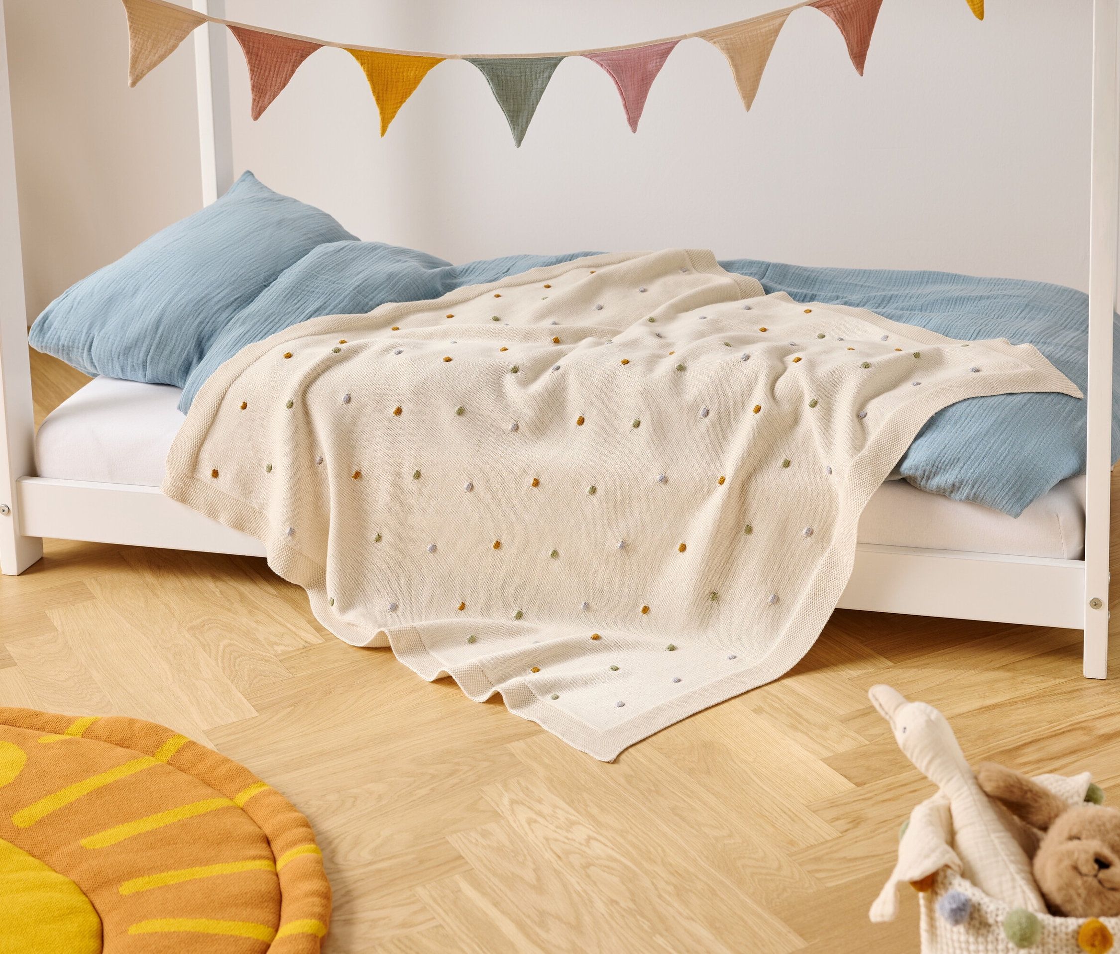 Łóżko dziecięce z niebieskimi poduszkami i kremowym kocem w kolorowe kropki stoi na drewnianej podłodze. Nad łóżkiem wisi kolorowy baner.