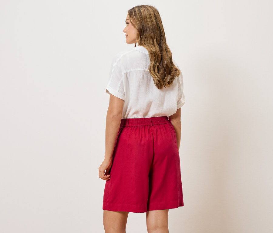 Widok z tyłu kobiety o falowanych włosach, ubranej w białą bluzkę i czerwone szorty.