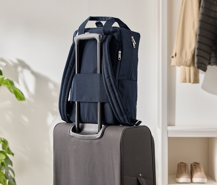 Granatowy plecak na szarym walizce na kółkach, obok roślina doniczkowa i szafa.