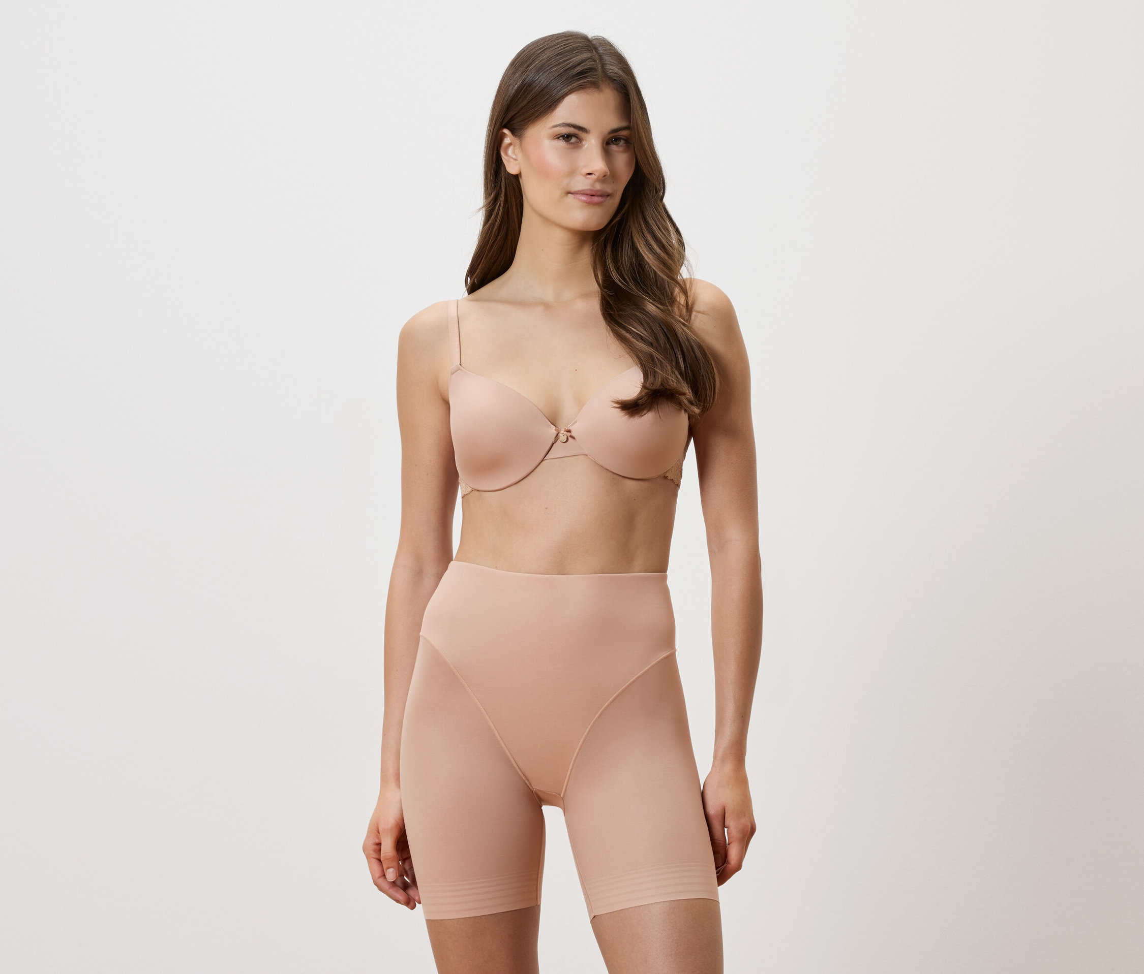 Kobieta ma na sobie beżowy biustonosz i modelujące szorty na białym tle.