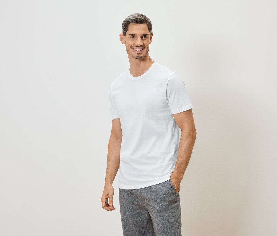 Uśmiechnięty mężczyzna z ręką w kieszeni, ubrany w biały T-shirt i szare szorty na białym tle.