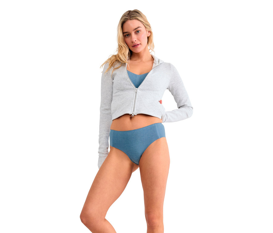 Kobieta z blond włosami stoi na białym tle, ubrana w jasnoszarą, krótką bluzę z kapturem na zamek, niebieski biustonosz typu bralette i niebieskie prążkowane figi. Jej ręce są za plecami.
