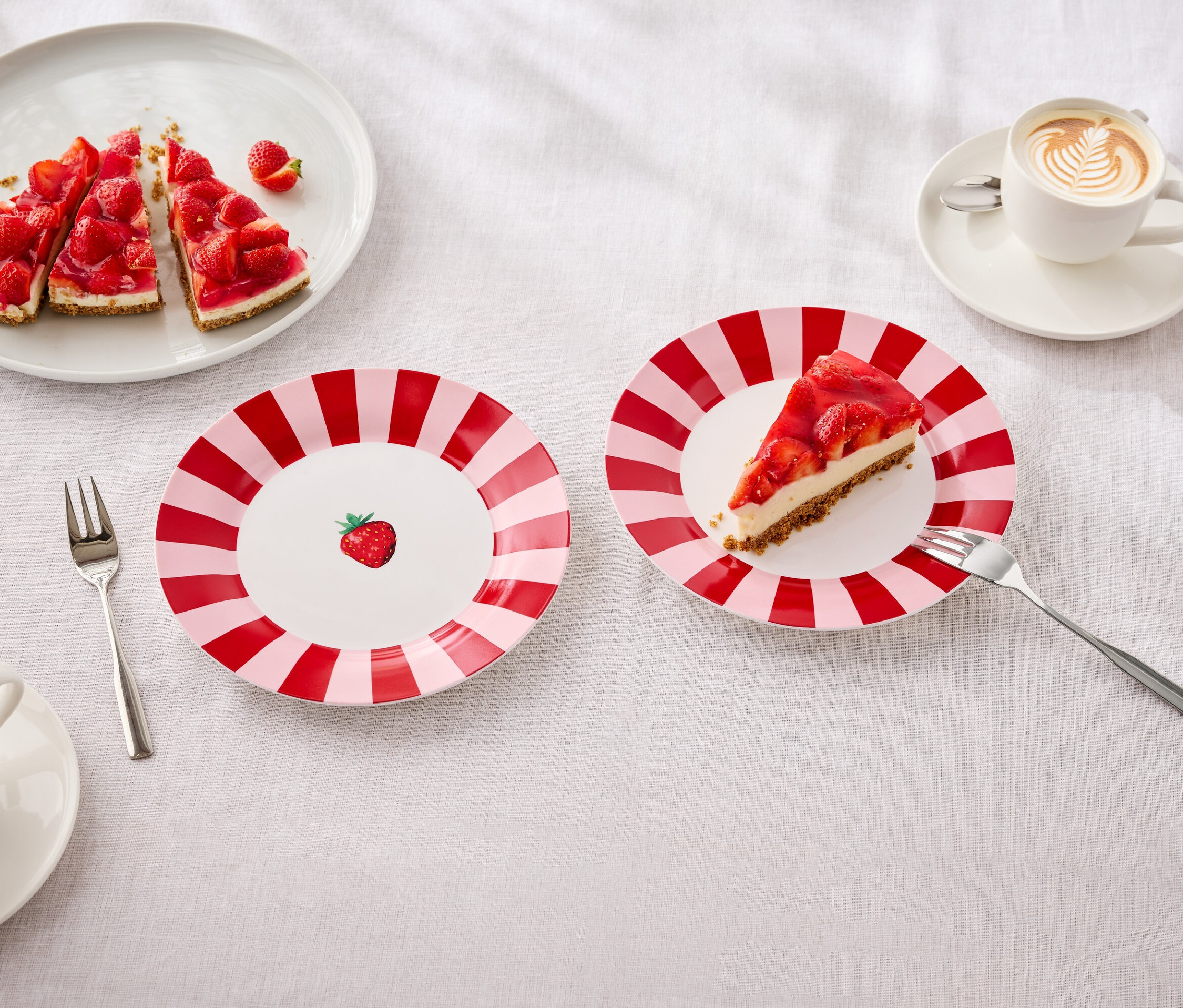 Na stole leżą talerze z ciastem truskawkowym, widelce i filiżanki z latte art.