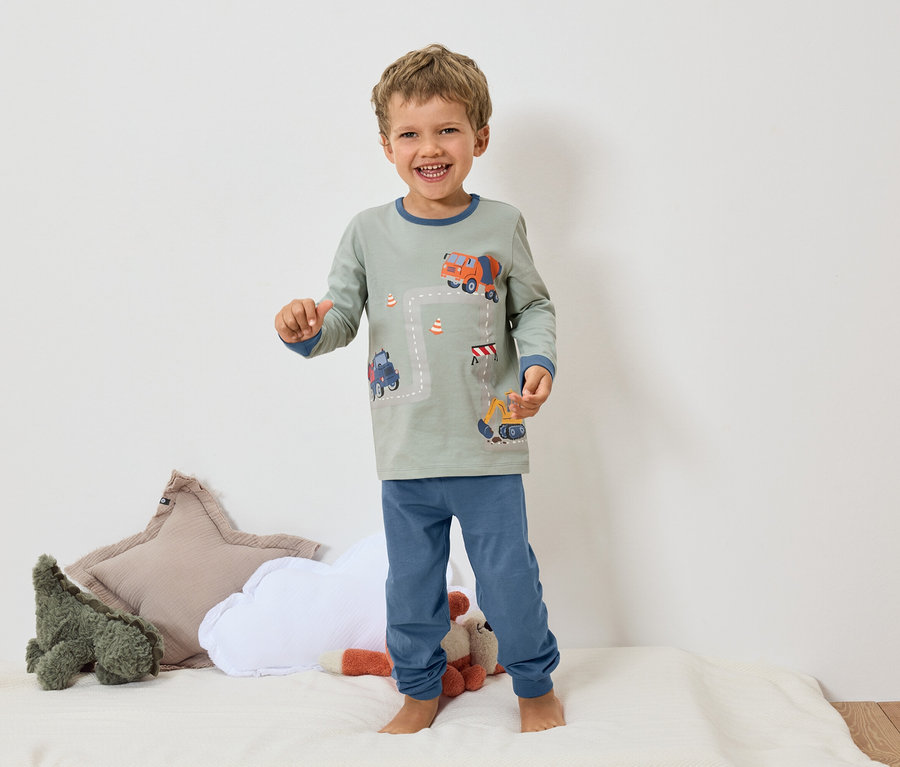 Uśmiechnięty chłopiec stoi boso na białej powierzchni, ubrany w szarą piżamę z niebieskimi mankietami i motywem koparki i betoniarki.