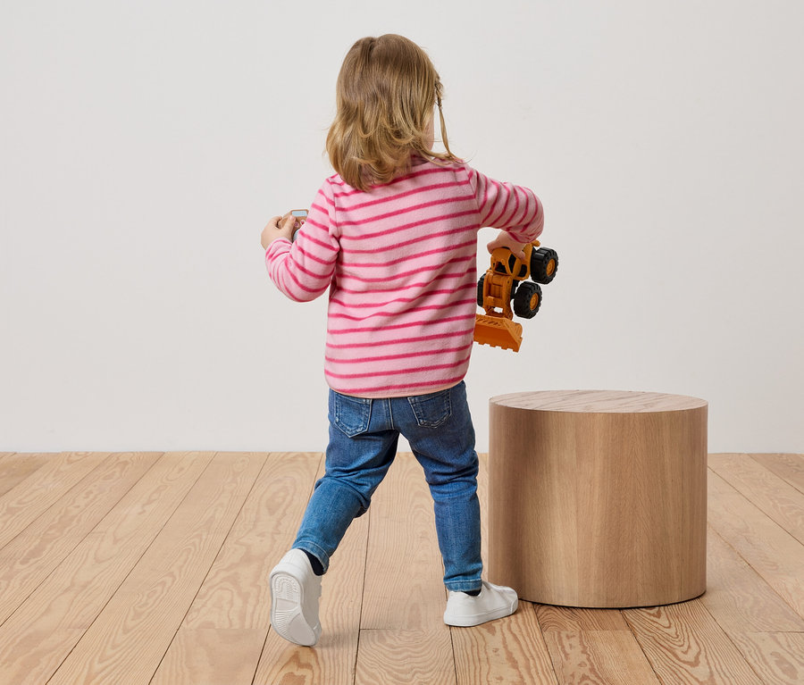 Dziecko z blond włosami stoi tyłem do widza i trzyma zabawkę koparki w ręku.