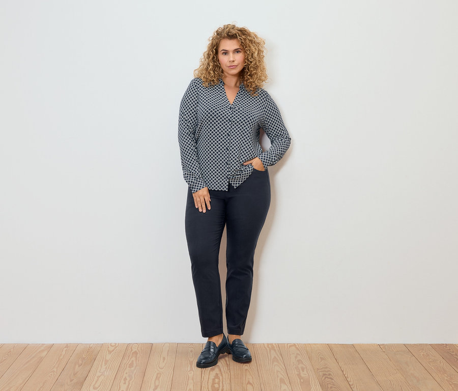 Kobieta w bluzce we wzory i spodniach z bengaliny pozuje na tle ściany.