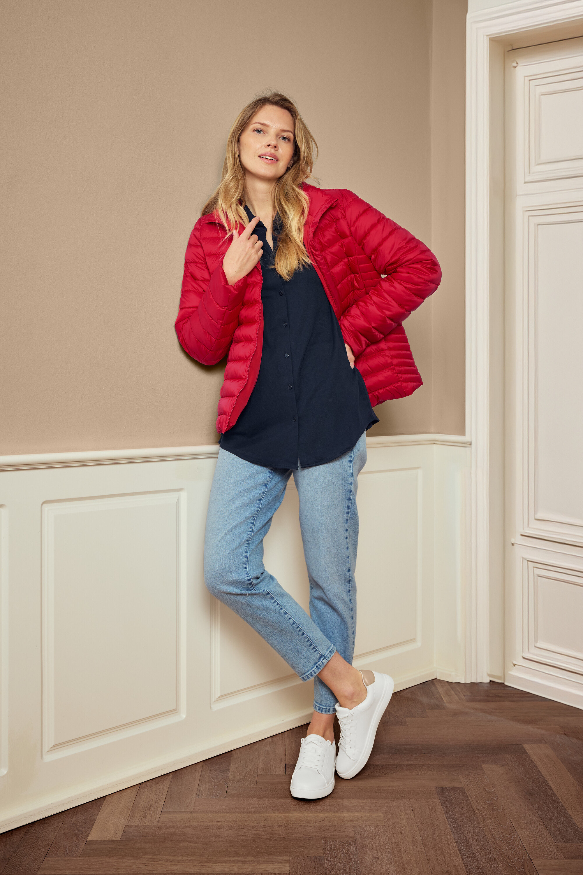 Kobieta pozuje w czerwonej kurtce, niebieskiej koszuli, dżinsach i białych butach sportowych, nosi kolczyki 925 Silber Creolen Hexagon.
