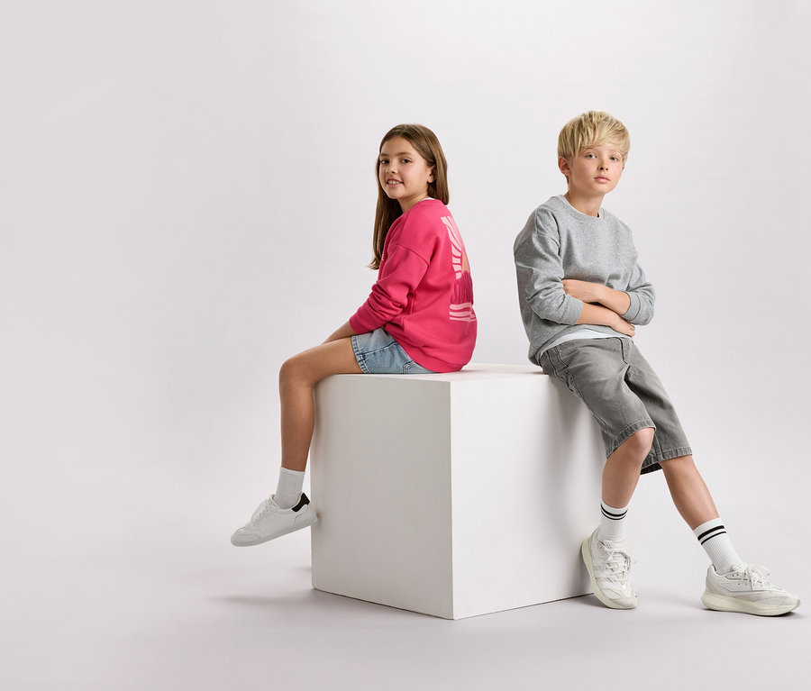 Dziewczynka z brązowymi włosami w różowej bluzie i dżinsowych szortach siedzi obok chłopca z blond włosami w szarej bluzie i szortach na białym sześcianie.