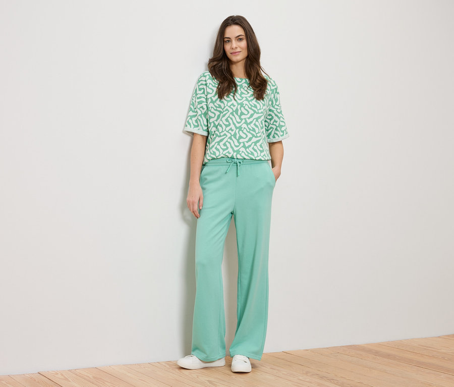 Kobieta opiera się o ścianę, ubrana w zieloną bluzkę we wzory i zielone spodnie.