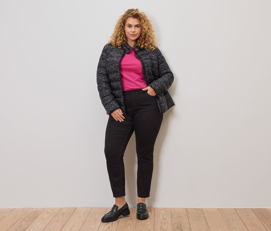 Kobieta o kręconych włosach ma na sobie szaro-czarną, pikowaną kurtkę nałożoną na różowy top, połączoną z czarnymi dżinsami i mokasynami.