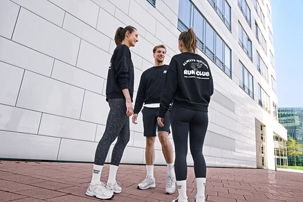 Trzy osoby, dwie kobiety i mężczyzna, stoją uśmiechnięte i rozmawiają. Wszyscy noszą czarne bluzy i białe skarpety tenisowe Coffee Crew unisex. Mężczyzna ma na sobie czarne szorty sportowe. Jedna kobieta nosi szaro-czarne legginsy sportowe „ActivePower”, druga czarne legginsy sportowe „ActivePower”.