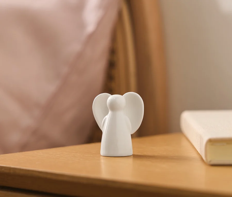 Biała ceramiczna figurka anioła, widziana od tyłu, stoi na drewnianym stoliku nocnym, z rozmytą różową poduszką i jasną książką w tle.