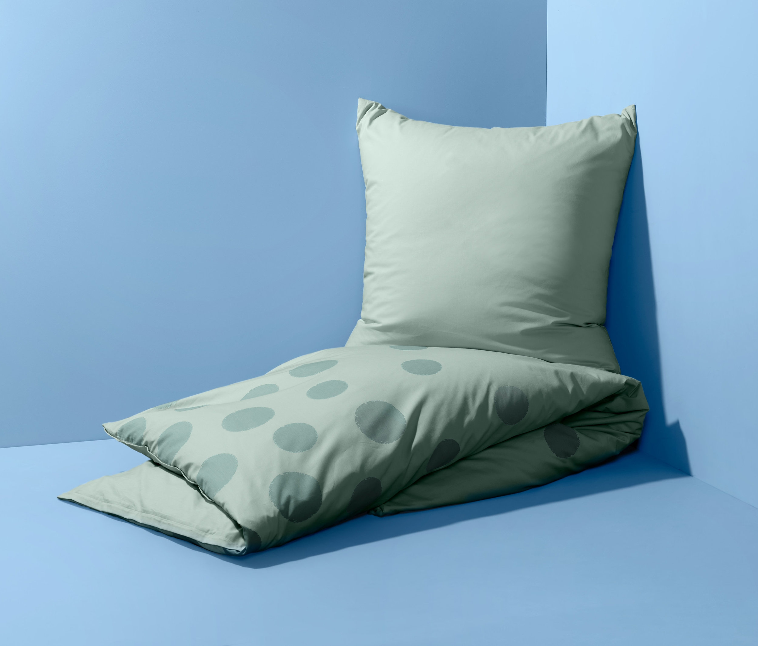 Jasnozielona, dwustronna pościel z perkalu w standardowym rozmiarze, w kropki, leży na podłodze przy ścianie.