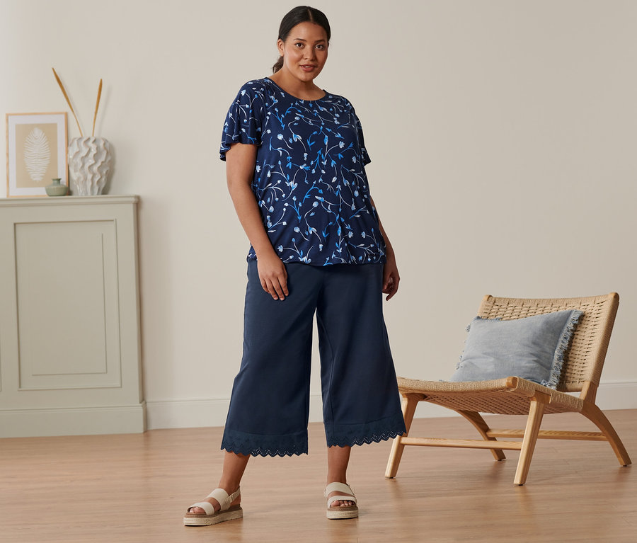 Kobieta stoi i ma na sobie granatową bluzkę z rękawami nietoperzowymi, kuloty z haftem i sandały skórzane na platformie.
