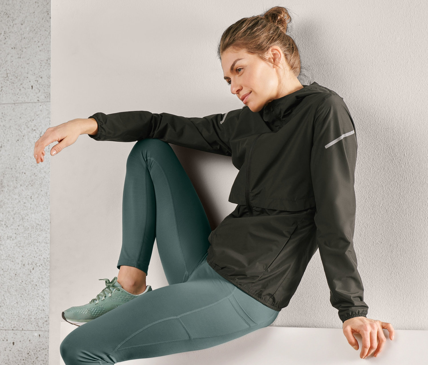 Kobieta siedzi na krawędzi, ubrana w czarną wiatroszczelną kurtkę do biegania, zielone legginsy i buty sportowe.
