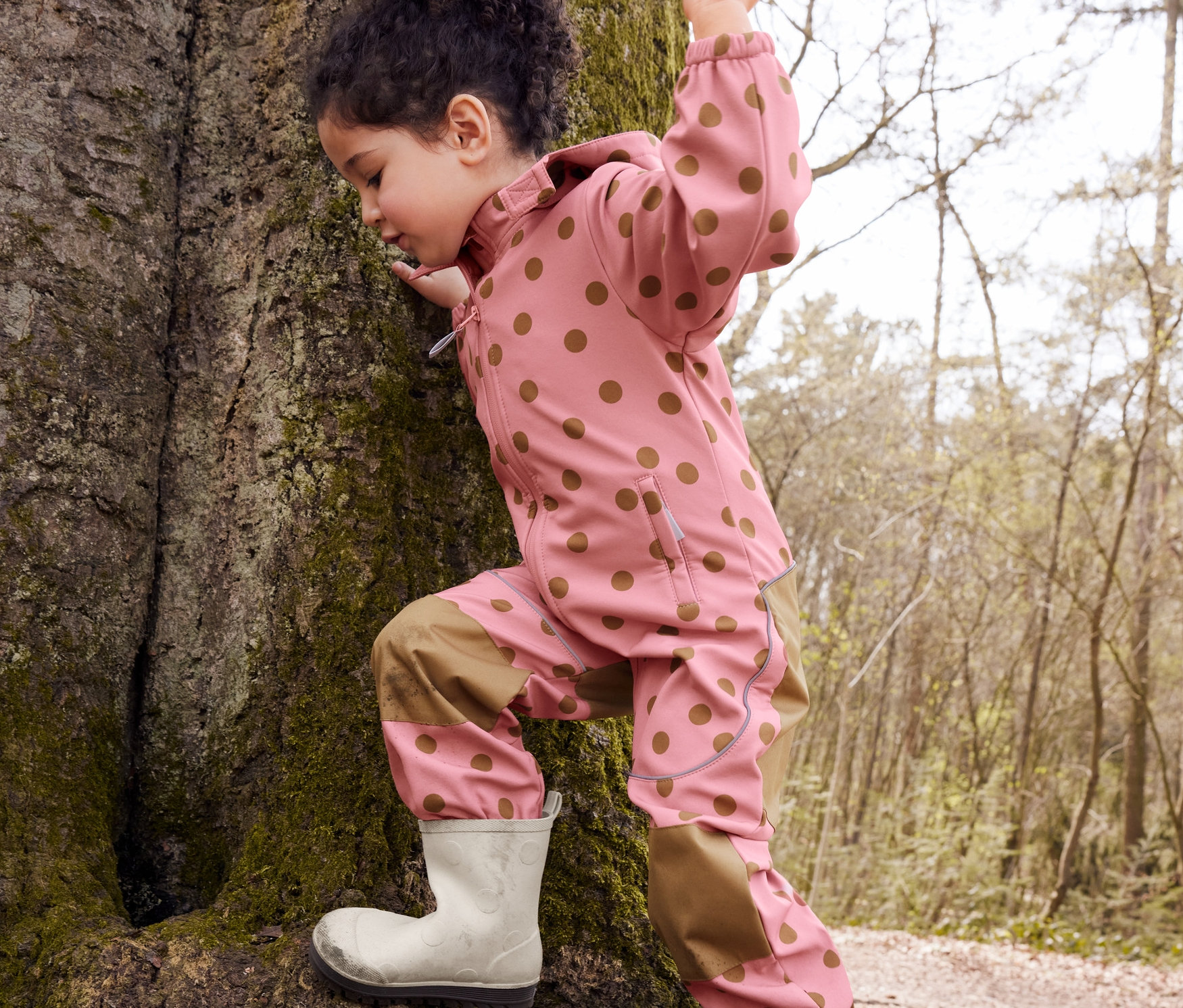 Dziewczynka wspina się na drzewo w jednoczęściowym kombinezonie softshell w kropki i kaloszach.