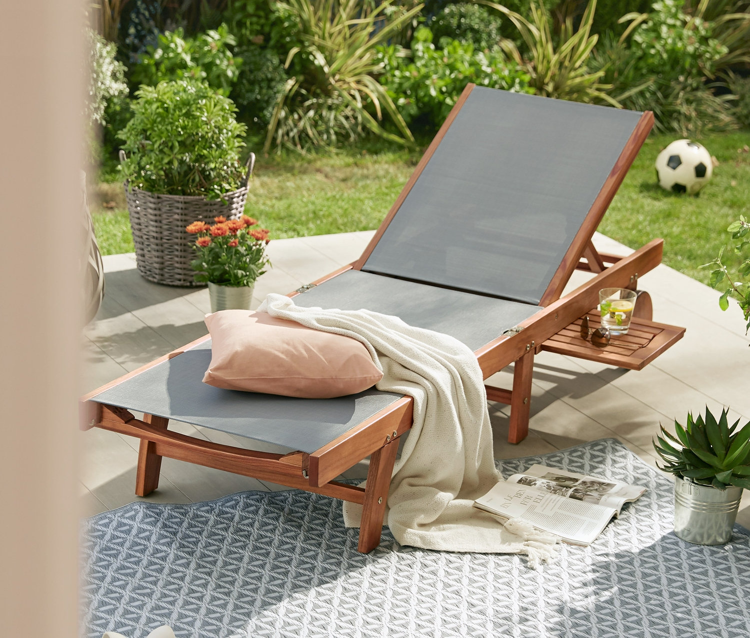 Składany leżak ogrodowy z poszyciem tekstylnym stoi na dywanie ogrodowym. W tle fontanna ogrodowa z oświetleniem LED.