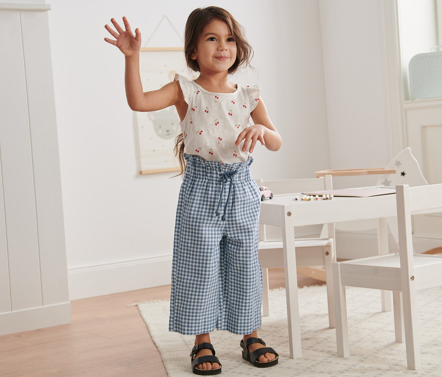 Dziewczyna macha i stoi w pokoju. Ma na sobie top z wisienkami i kuloty z lnem.