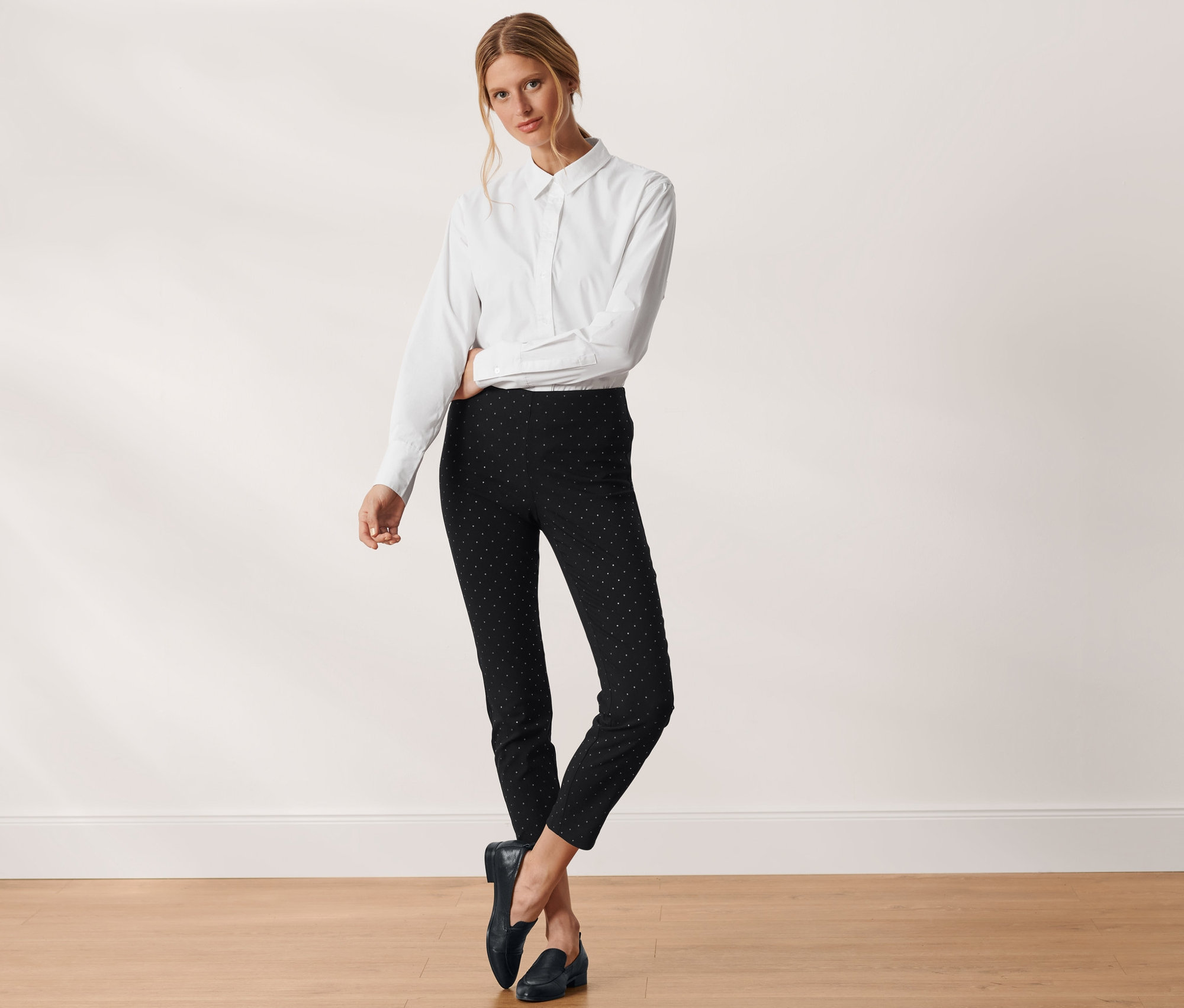Kobieta pozuje w białej koszuli i czarnych spodniach ze stretchu w kropki.