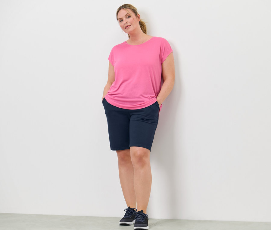 Kobieta stoi z rękami w kieszeniach szortów. Ma na sobie różową koszulkę sportową.