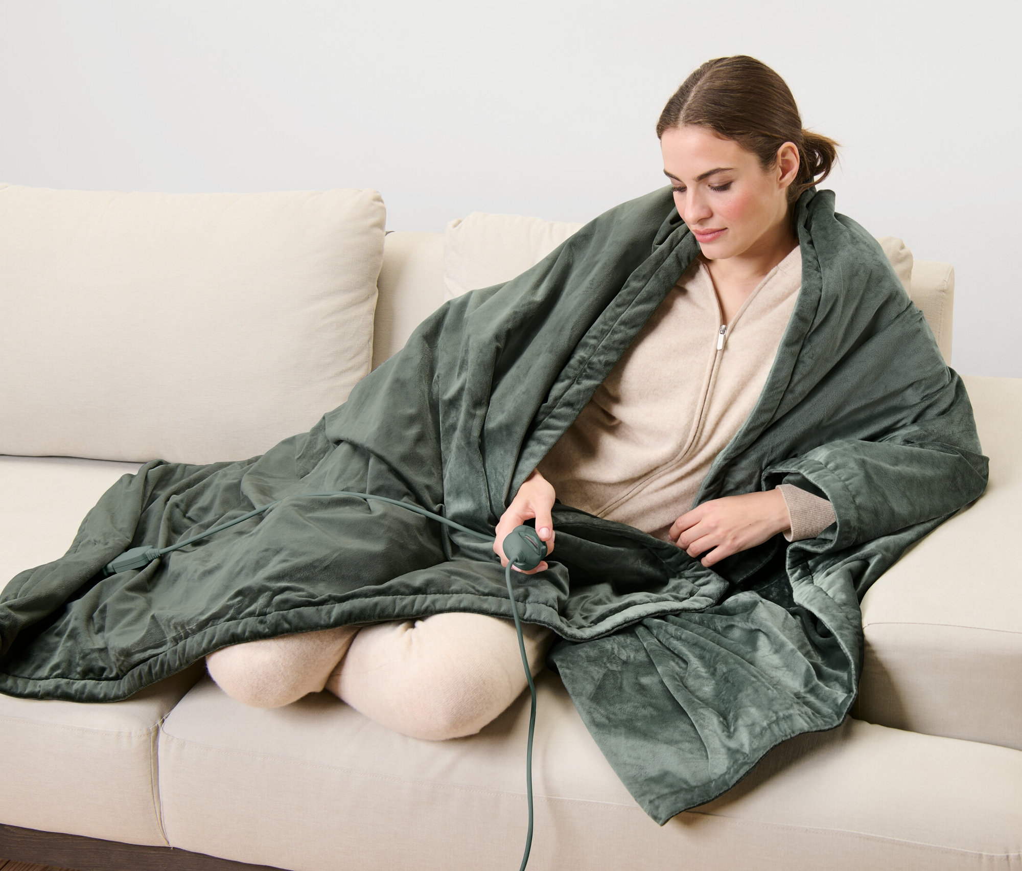 Kobieta siedzi na kanapie, przykryta kocem elektrycznym i trzyma pilot.
