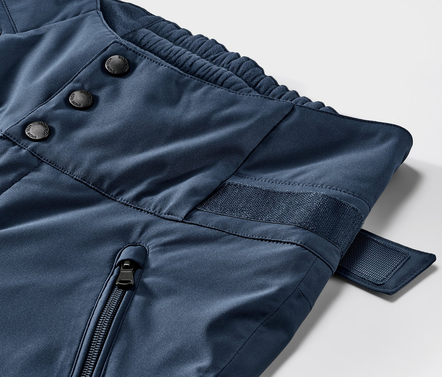 Zbliżenie na niebieskie spodnie narciarskie z wysokim stanem. Widoczne detale: guziki, zamek błyskawiczny i rzepy.