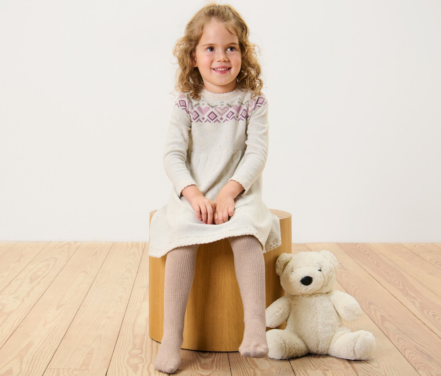 Dziewczynka siedzi na drewnianym stołku, ubrana w sukienkę i Rajstopy z wełną z certyfikatem RWS.