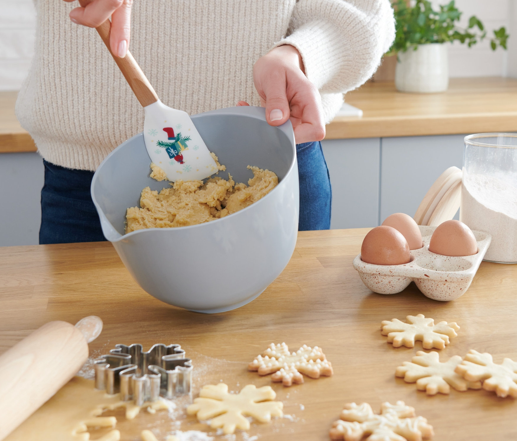 Kobieta miesza ciasto w misce za pomocą skrobaka do ciasta. Na stole leżą wykrojone kształty płatków śniegu, wałek i jajka.